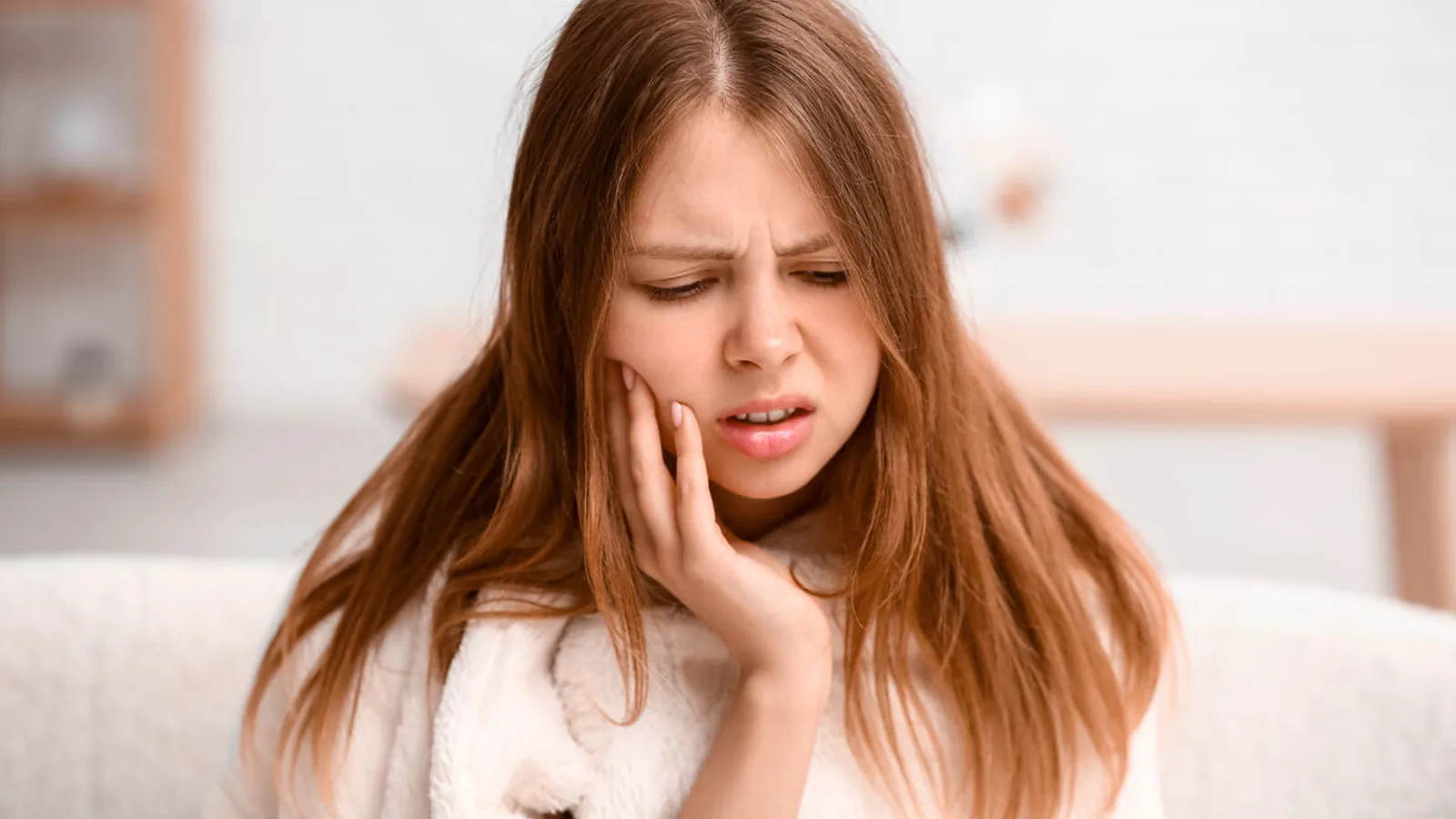 Jeune femme assise dans son salon, la main pressée contre la joue, le visage crispé